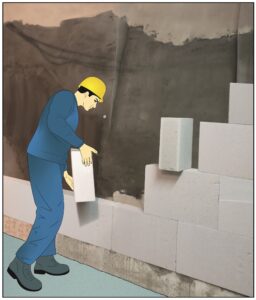 Brugger AG Mann Steht mit Gelbem Helm und blauer Arbeitskleidung vor Kellerwand und Isoliert die Kellerwände